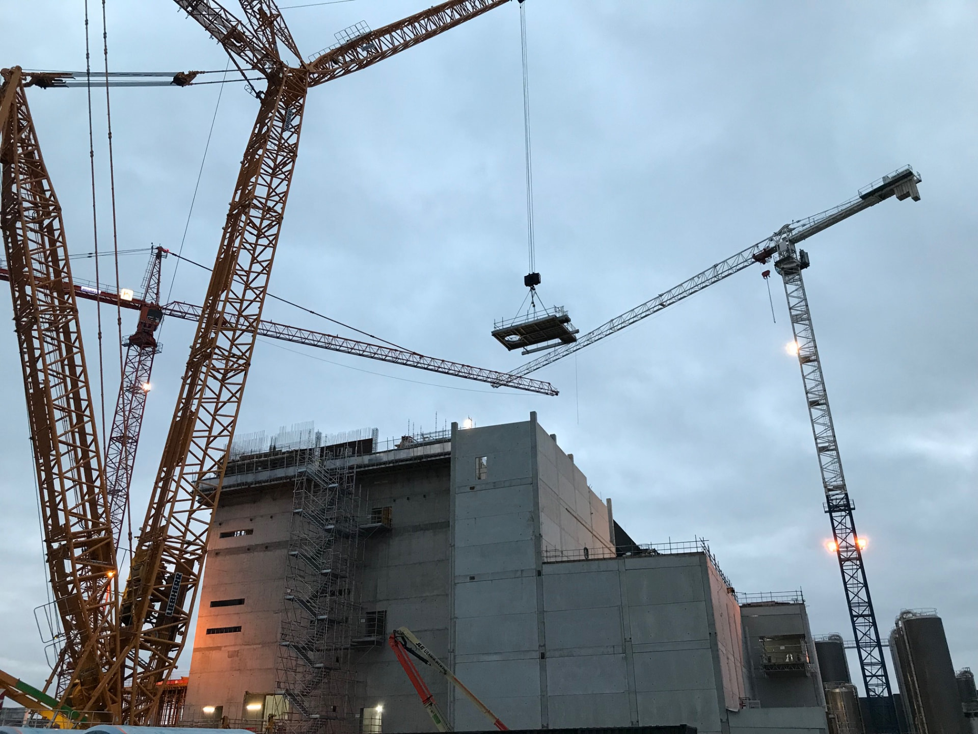 Tower cranes lifting a steel platform module onto a large industrial facility under construction at dusk